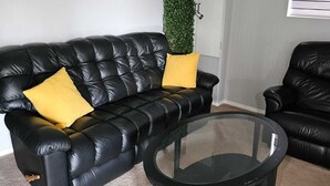A living room with a black leather sofa, two yellow cushions, a glass coffee table, and a green vertical plant.