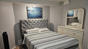 A bedroom with a tufted headboard, a bed with striped bedding, a dresser with drawers, a mirror, and a picture on the wall.