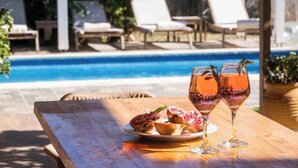A wooden table with a plate of food and two glasses of a pink beverage, a pool, and lounge chairs in the background. - Rigilis - Two Bedroom Villa, Sleeps 4 (Nydri)
