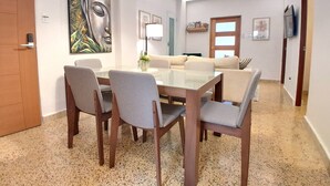 A modern dining area with a glass table, wooden chairs, a sofa, and a ceiling light.