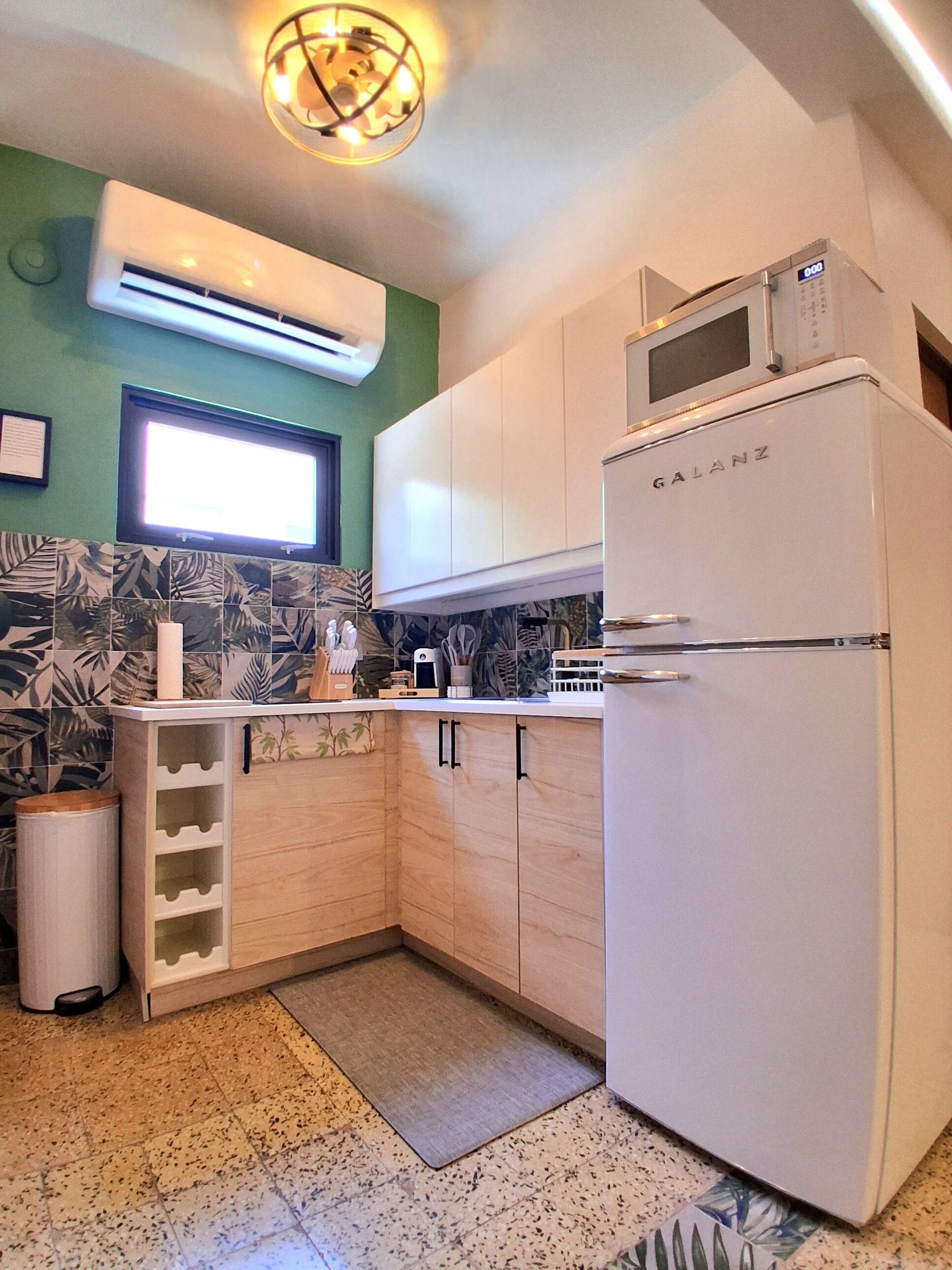 A compact kitchen with a white refrigerator, a microwave, and a small sink. There is a window above the sink and a wall-mounted air conditioner.