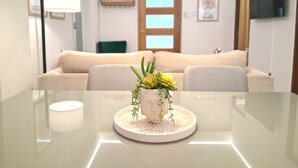 A modern living room with a glass table, a beige sofa, a decorative plant centerpiece, and a flat-screen TV mounted on the wall.
