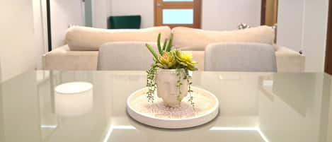 A modern living room with a glass table, a beige sofa, a decorative plant centerpiece, and a flat-screen TV mounted on the wall.