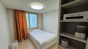 A compact bedroom with a single bed, a window with curtains, a ceiling light, a heater, and a shelf with a kettle and a container.