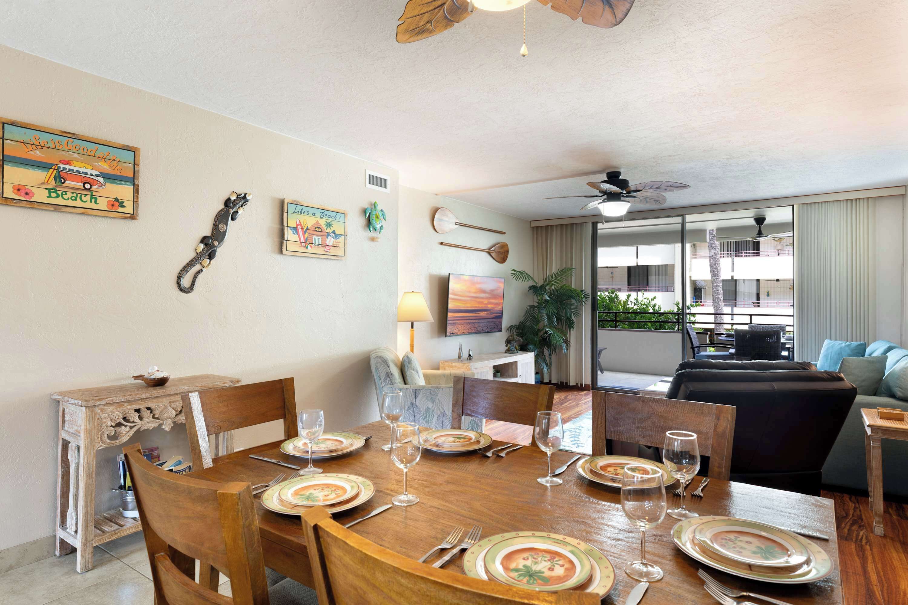 Un coin repas avec une table en bois dressée pour quatre personnes, entourée de chaises. Des éléments décoratifs ornent les murs et un ventilateur de plafond est installé au-dessus.