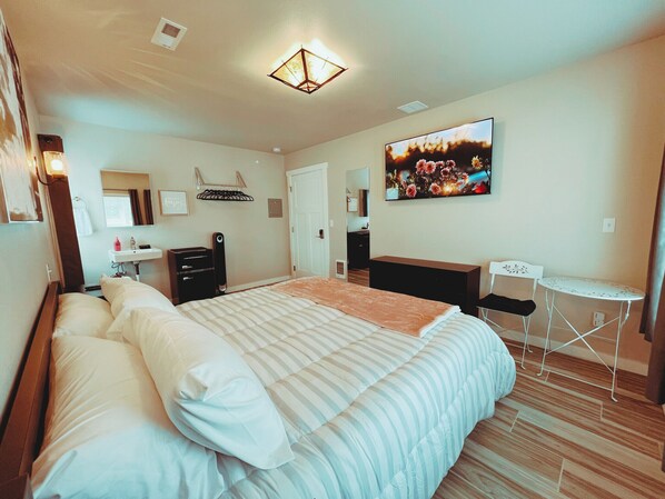 A hotel room with a large bed, a desk, a chair, a small table, a mirror, a sink, and a television.