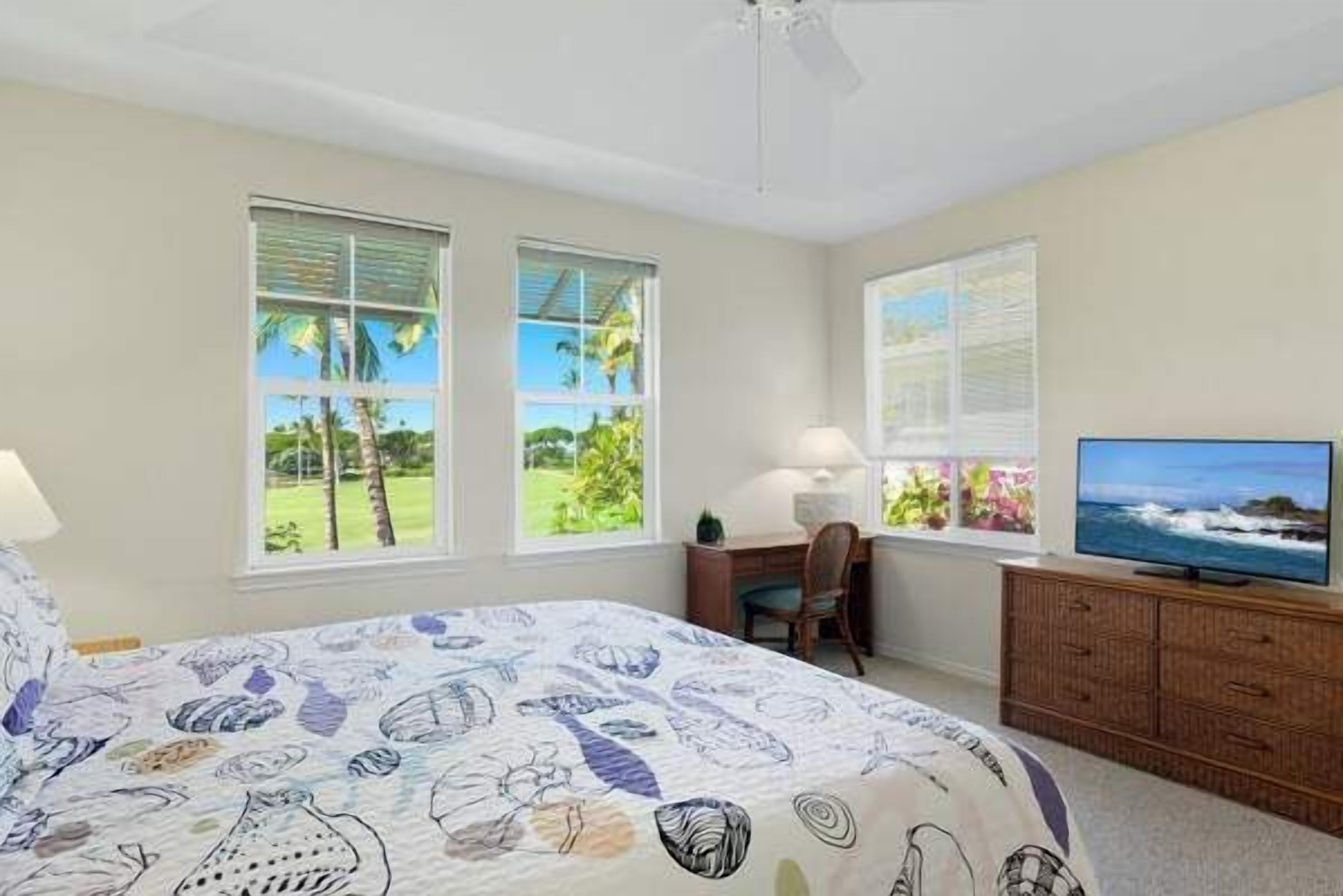 A bedroom with a bed, a desk, a chair, a television, and windows with a view of greenery.