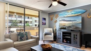 A living room with a sofa, armchair, and TV, a ceiling fan, and a large window overlooking a balcony. - Magic Sands Unit 111: Beach and Saltwater Pool (Kailua-Kona)