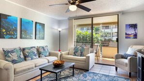 A living room with a sofa, coffee table, and chairs, a ceiling fan, and artwork on the walls. - Magic Sands Unit 111: Beach and Saltwater Pool (Kailua-Kona)