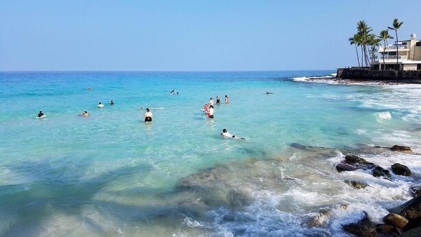 A beach with clear turquoise water, people swimming and surfing, and a building with palm trees on the right. - Magic Sands Unit 111: Beach and Saltwater Pool (Kailua-Kona)