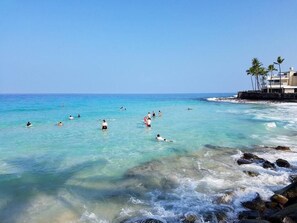 A beach with clear turquoise water, people swimming and surfing, and a building with palm trees on the right. - Magic Sands Unit 111: Beach and Saltwater Pool (Kailua-Kona)