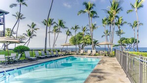 Une piscine entourée de chaises longues et de parasols, bordée de palmiers et offrant une vue sur l’océan.