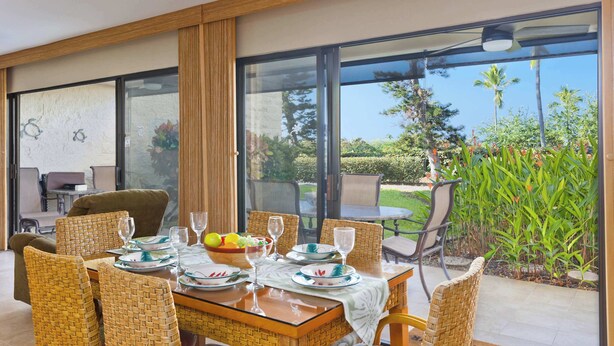 Un comedor con una mesa dispuesta para seis personas, rodeado de sillas de mimbre, y vistas al exterior con follaje y palmeras.