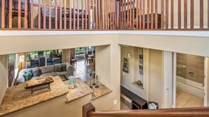 A spacious living area with a large sectional sofa, a coffee table, and a dining area with chairs. A staircase connects the two levels. - Hale Laki – Stunning Resort Condo, Close to Beach! (Waikoloa)