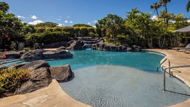 A pool with a waterfall feature surrounded by tropical vegetation. - Hale Laki – Stunning Resort Condo, Close to Beach! (Waikoloa)