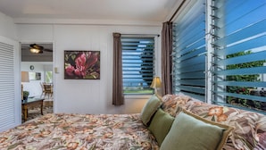 Een slaapkamer met een bloemenbedekking, uitzicht door een raam met jaloezieën, een bloemschilderij en een lamp op de nachtkastje.