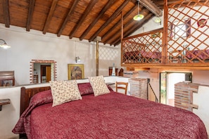 A bedroom with a wooden ceiling, a bed with a red bedspread, a wooden chair, and a brick wall.