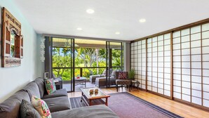 A modern living room with a sofa, chairs, and a coffee table. There is a sliding glass door leading to a balcony with a view of greenery.