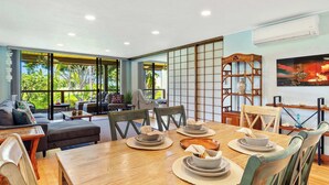 A modern living room with a large dining table set for a meal, a sofa, and a view of lush greenery outside.