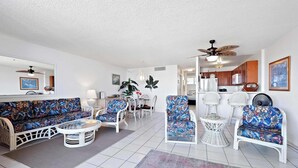 A living room with a sofa, chairs, a coffee table, and a dining table. There is a ceiling fan, a kitchen area in the background, and a large mirror on the wall. - Kolu Honu at Kona Ali'i- Stunning Beachfront Condo (Kailua-Kona)
