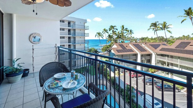 A balcony with a table set for two, a ceiling fan, and a view of the ocean and palm trees. - Kolu Honu at Kona Ali'i- Stunning Beachfront Condo (Kailua-Kona)