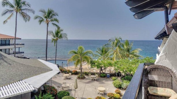 A balcony with a view of the ocean, palm trees, and outdoor seating area.