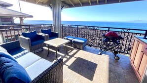 A covered outdoor seating area with wicker furniture and a grill, overlooking a rocky beach.