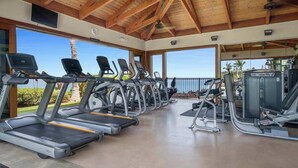 A covered gym area with treadmills, elliptical machines, and weight equipment, featuring large windows and a wooden ceiling.