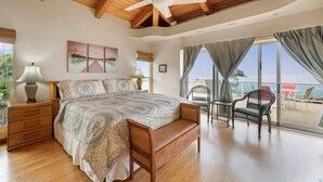 Une chambre avec un grand lit, une tête de lit en bois et un ventilateur de plafond. Il y a une fenêtre avec des rideaux et une vue sur l’extérieur.