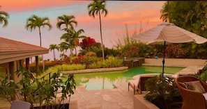 Zona de piscina con sillas de descanso y sombrillas, rodeada de palmeras y un cielo al atardecer.