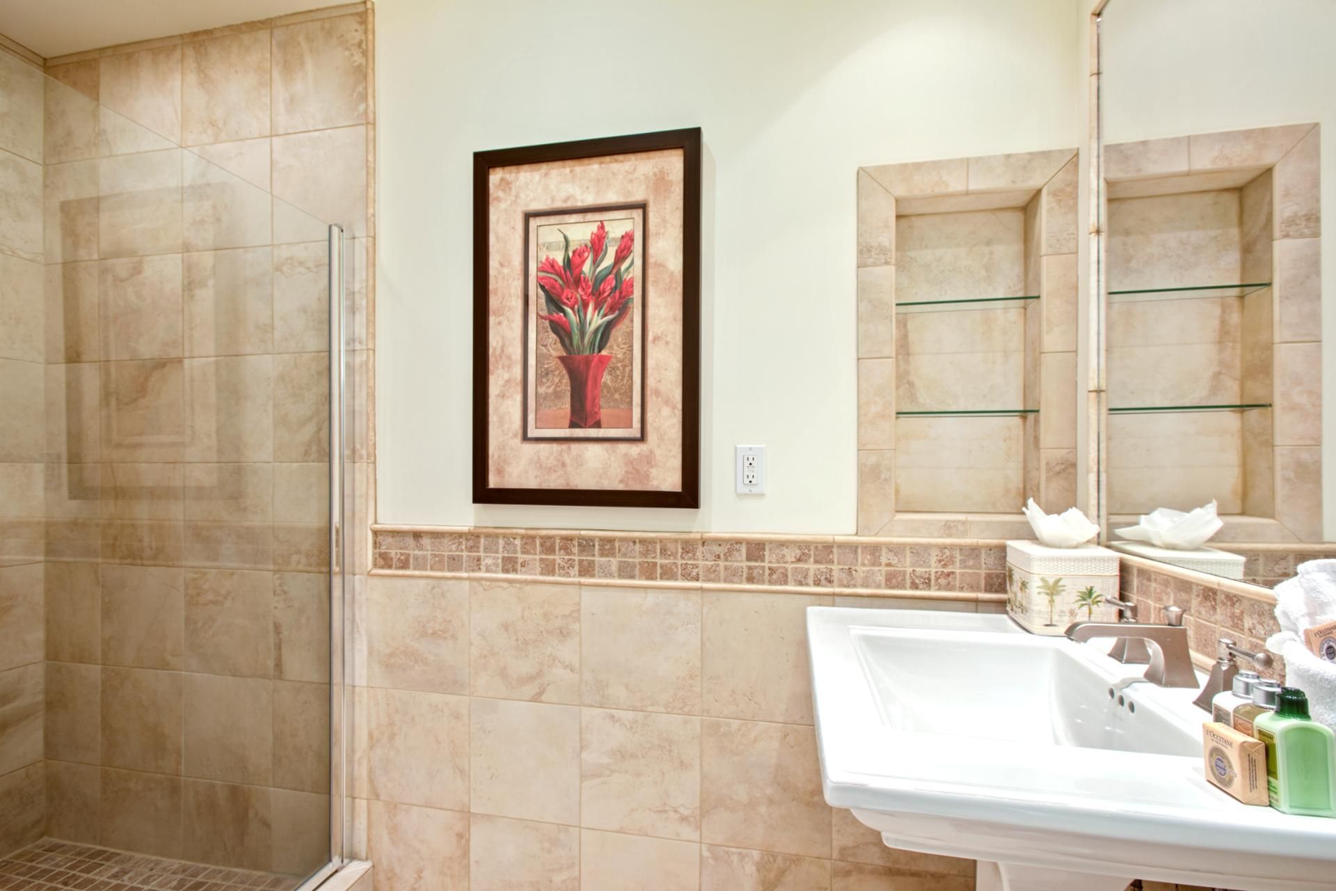 A bathroom with beige tiles, a framed picture of red flowers, a glass shower enclosure, and a sink with toiletries.