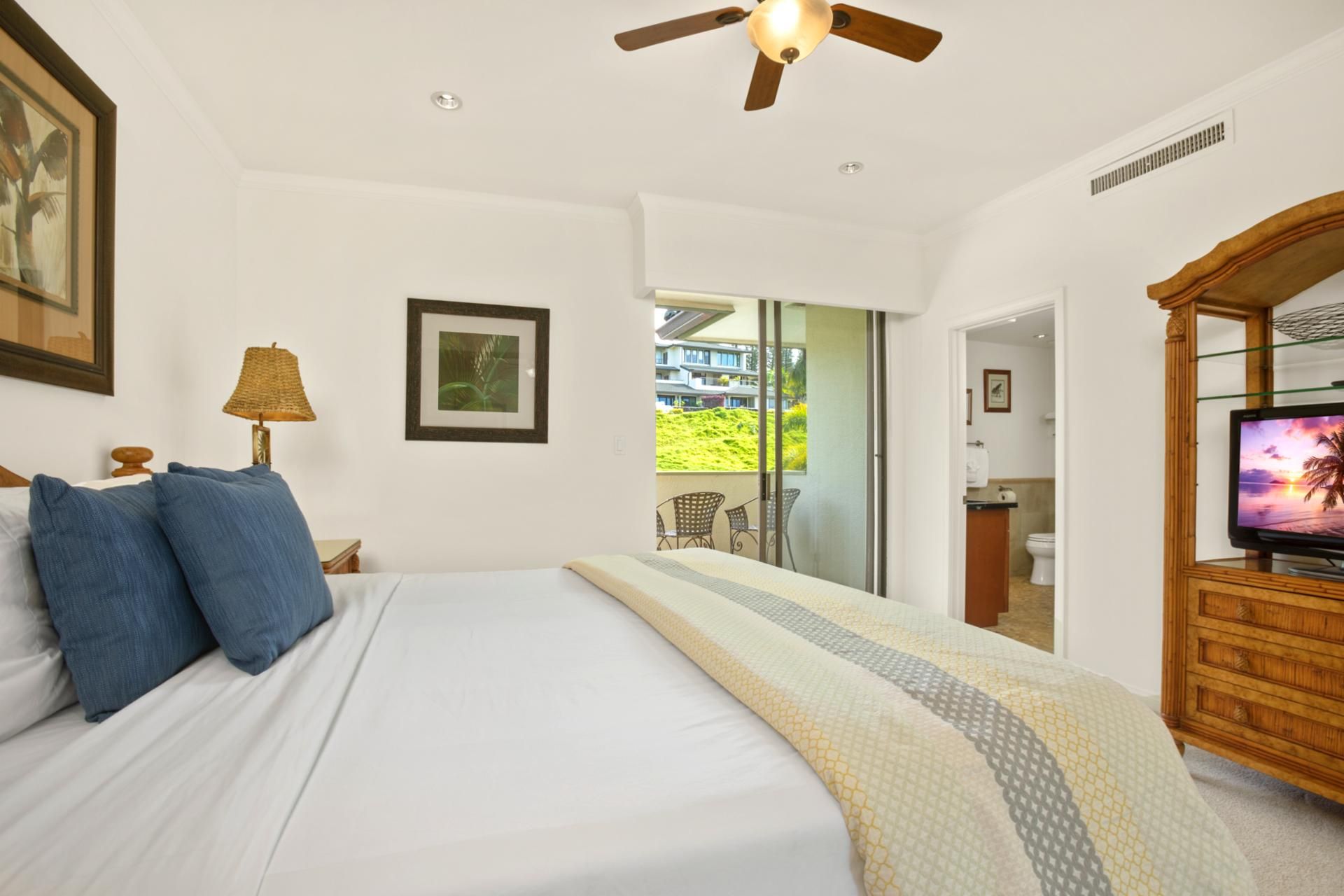 A bedroom with a bed, a ceiling fan, a TV, and a balcony with a view of buildings and greenery.