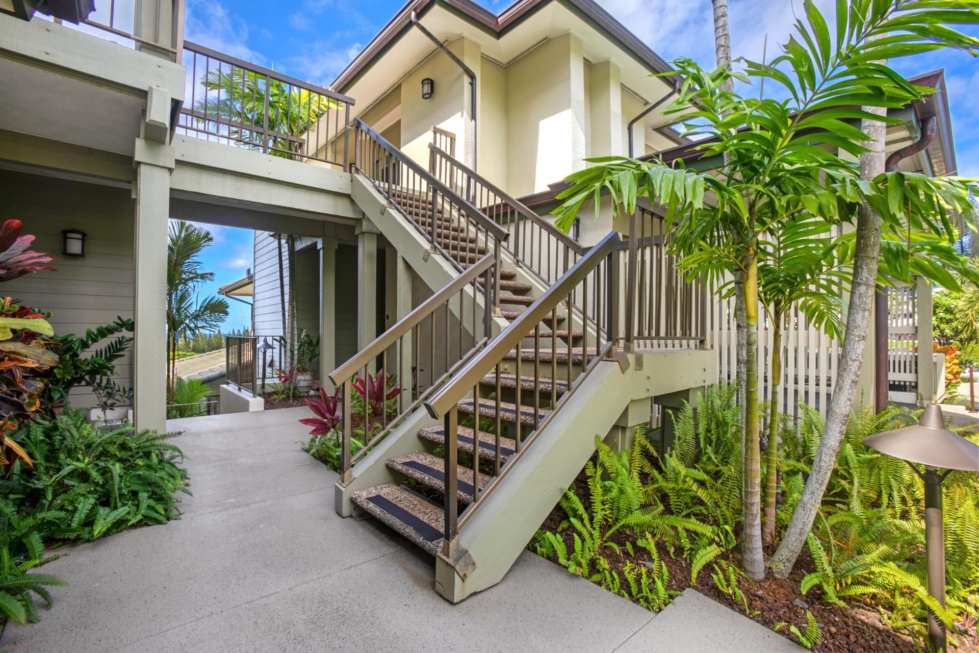 A two-story house with a staircase leading to the upper floor, surrounded by lush tropical plants.