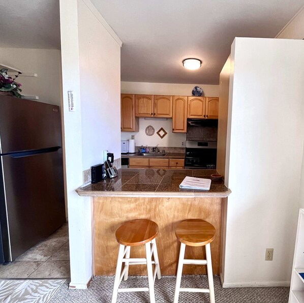 Una cocina compacta con armarios de madera, horno empotrado, refrigerador y dos taburetes.