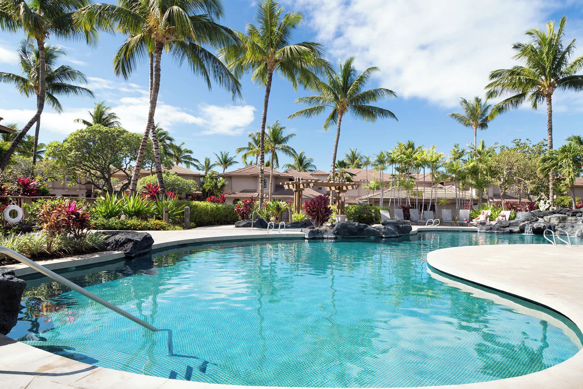 A resort pool with clear blue water, surrounded by palm trees and tropical plants.