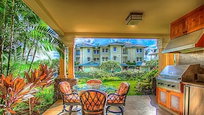 An outdoor kitchen area with a grill, surrounded by tropical plants and a view of a multi-story building. - Kolea 11D | Walk to the Beach (Waikoloa Beach Resort)