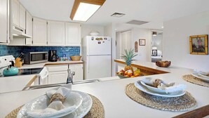 Una cocina moderna con armarios blancos, microondas, cafetera y refrigerador. Hay un frutero en la encimera y una zona de comedor con individuales y platos.