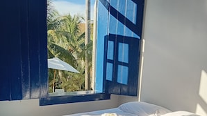 A room with a bed, a window with blue shutters, and a view of palm trees outside. - Pousada Usina Paraíso (Astolfo Dutra)