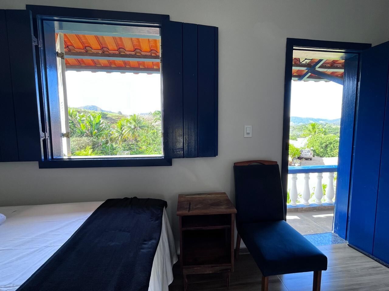 A room with a bed, a small wooden table, a bench, and a balcony with a view of greenery and hills.