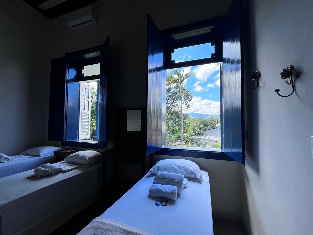 Une chambre avec deux lits, une fenêtre donnant sur un espace verdoyant et un luminaire fixé au mur.