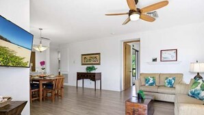 Amplia sala de estar con suelo de madera, ventilador de techo, mesa de comedor con sillas, sofá con cojines y un cuadro en la pared.