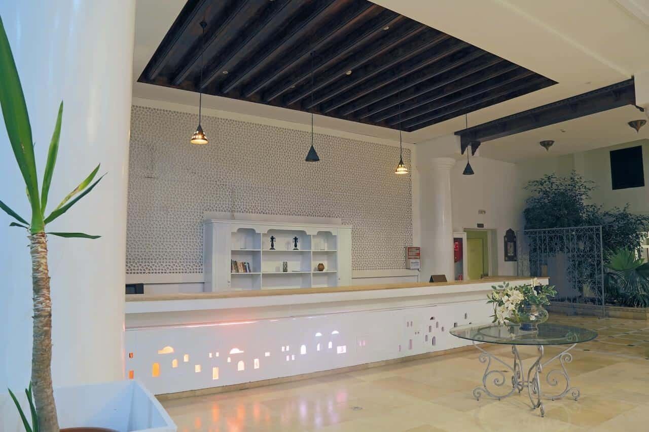 Un área de recepción con diseño moderno, que presenta un mostrador blanco, un muro con diseño y luminarias colgantes.