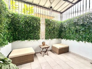 A covered patio with a seating area, a small table, and lush green foliage on the walls.