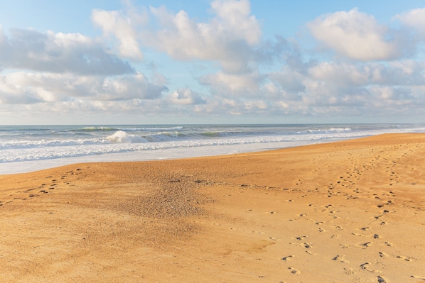 Ein sandiger Strand mit Fußspuren, die zum Wasser führen, ein klarer Himmel und ansteigende Wellen.