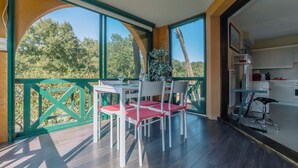 A dining area with a table and chairs, a view of trees, and a kitchen area with cabinets and appliances. - Le Landais - Wifi - Loggia - Plage (Tarnos)