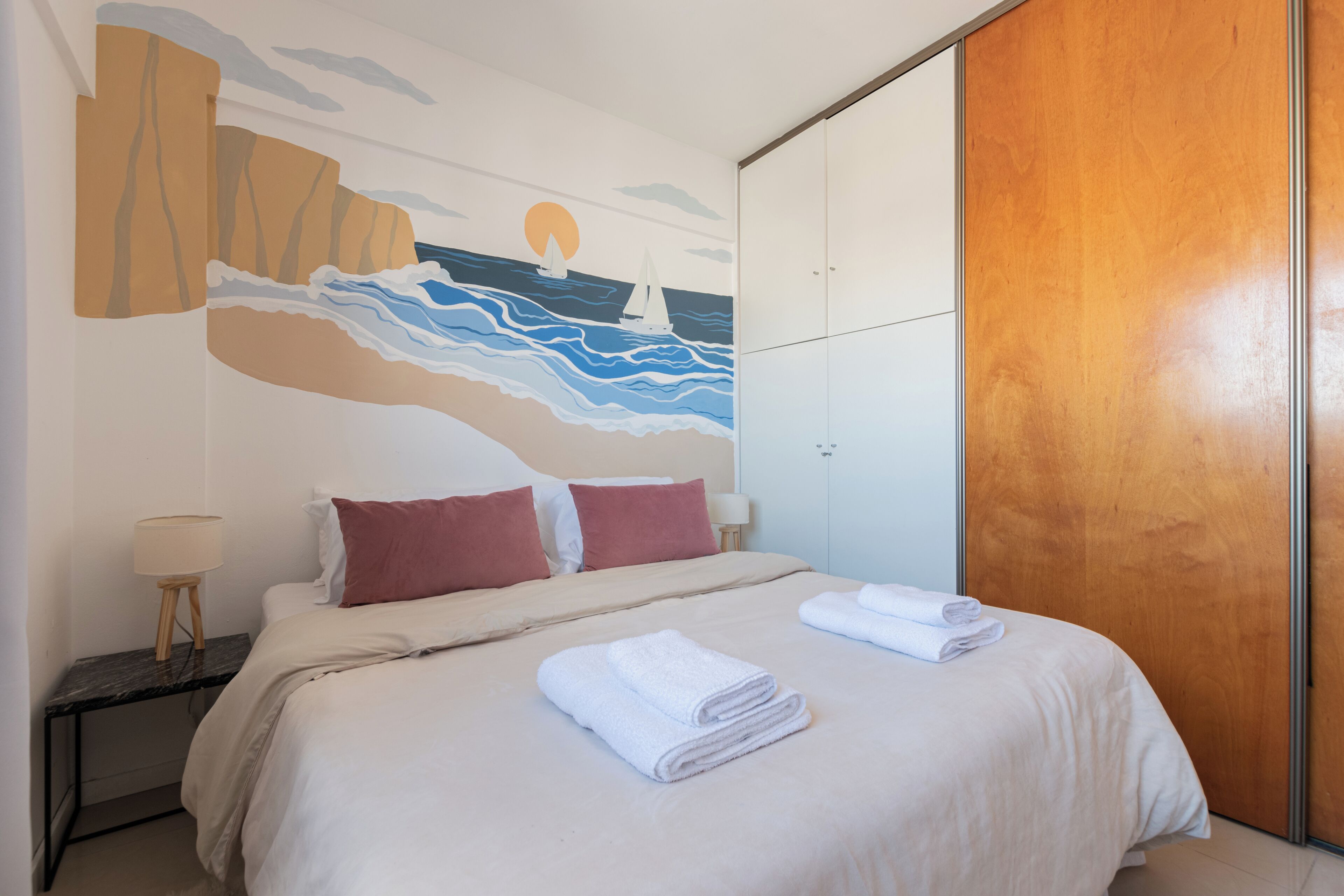 Una cama bien hecha con dos almohadas, un mural en la pared que muestra un atardecer sobre el mar y un armario de madera.