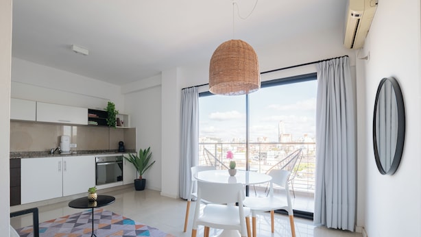 Una cocina moderna con armarios blancos, un comedor con mesa y sillas blancas, una lámpara colgante de mimbre y vistas de la ciudad a través de grandes ventanales.