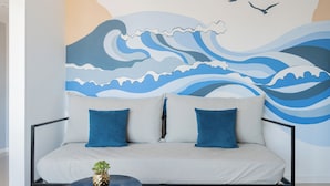Una sala moderna con un sofá, dos cojines azules, una mesita redonda con una planta y un mural en la pared que muestra olas y pájaros.