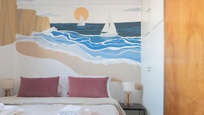 Habitación de hotel con cama, dos almohadas, mesita de noche con lámpara y un mural en la pared que muestra un velero en el mar.