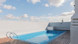 Una piscina en la azotea con un deck de madera, una pared blanca y cielo despejado con nubes.
